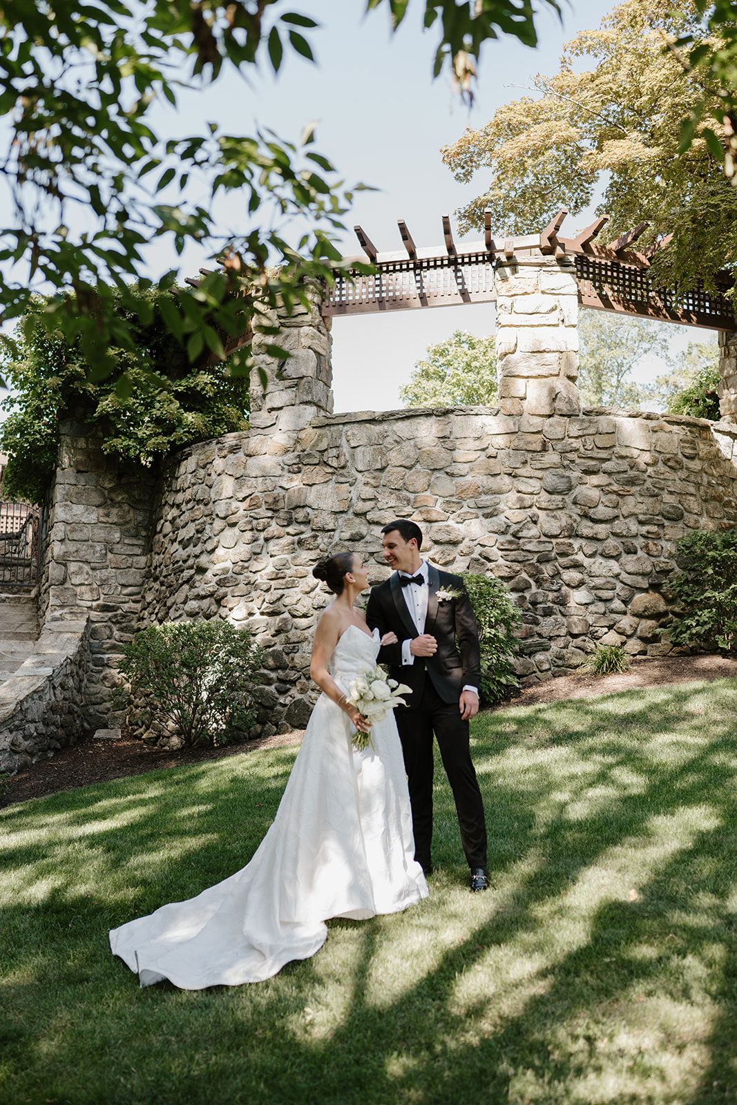 Bride and groom standing arm-in-arm by a stone wall, smiling lovingly at each other under dappled sunlight.